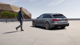 Mattgrauer Audi RS 6 Avant performance auf Platz, Mann im Anzug nähert sich, modernes Gebäude mit goldfarbener Fassade im Hintergrund.