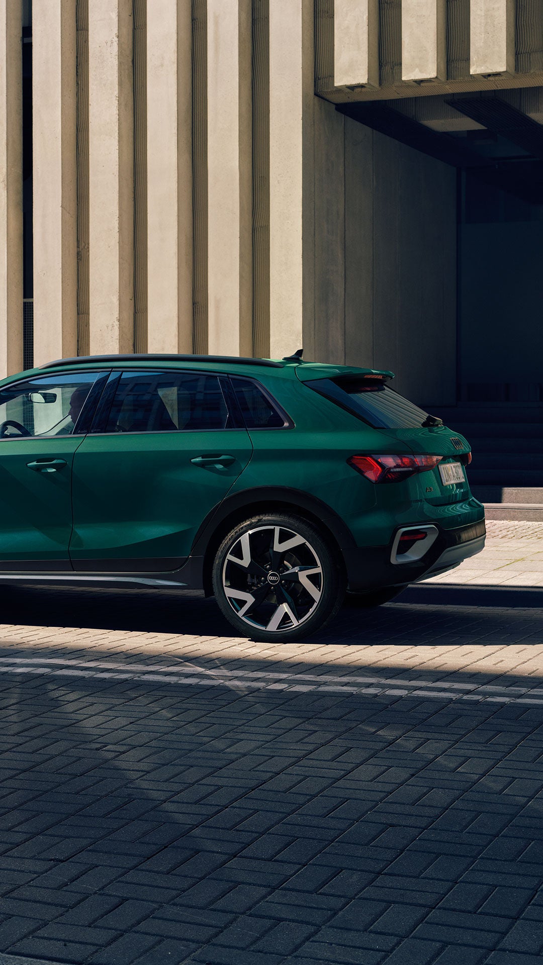 Grüner Audi A3 allstreet von der Seite vor moderner Betonfassade, Frau in gelber Kleidung geht auf dem Gehweg.