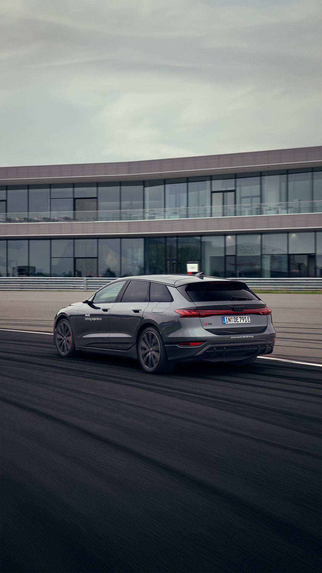 Ein grauer Audi S6 Avant e-tron mit der Aufschrift „Audi driving experience“ fährt auf einer Teststrecke vor einem modernen Gebäude. 