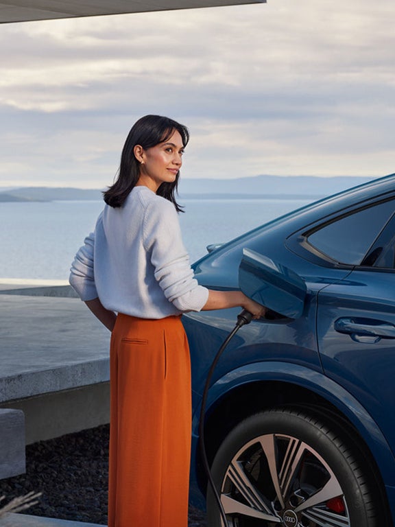 Frau mit heller Bluse und orangem Rock lädt blauen Audi SQ6 Sportback e-tron an einer Wandladestation.