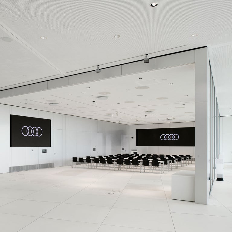 Großer Konferenzraum mit zwei Bildschirmen mit Audi Logo und zahlreichen schwarzen Stühlen in Reihenanordnung.