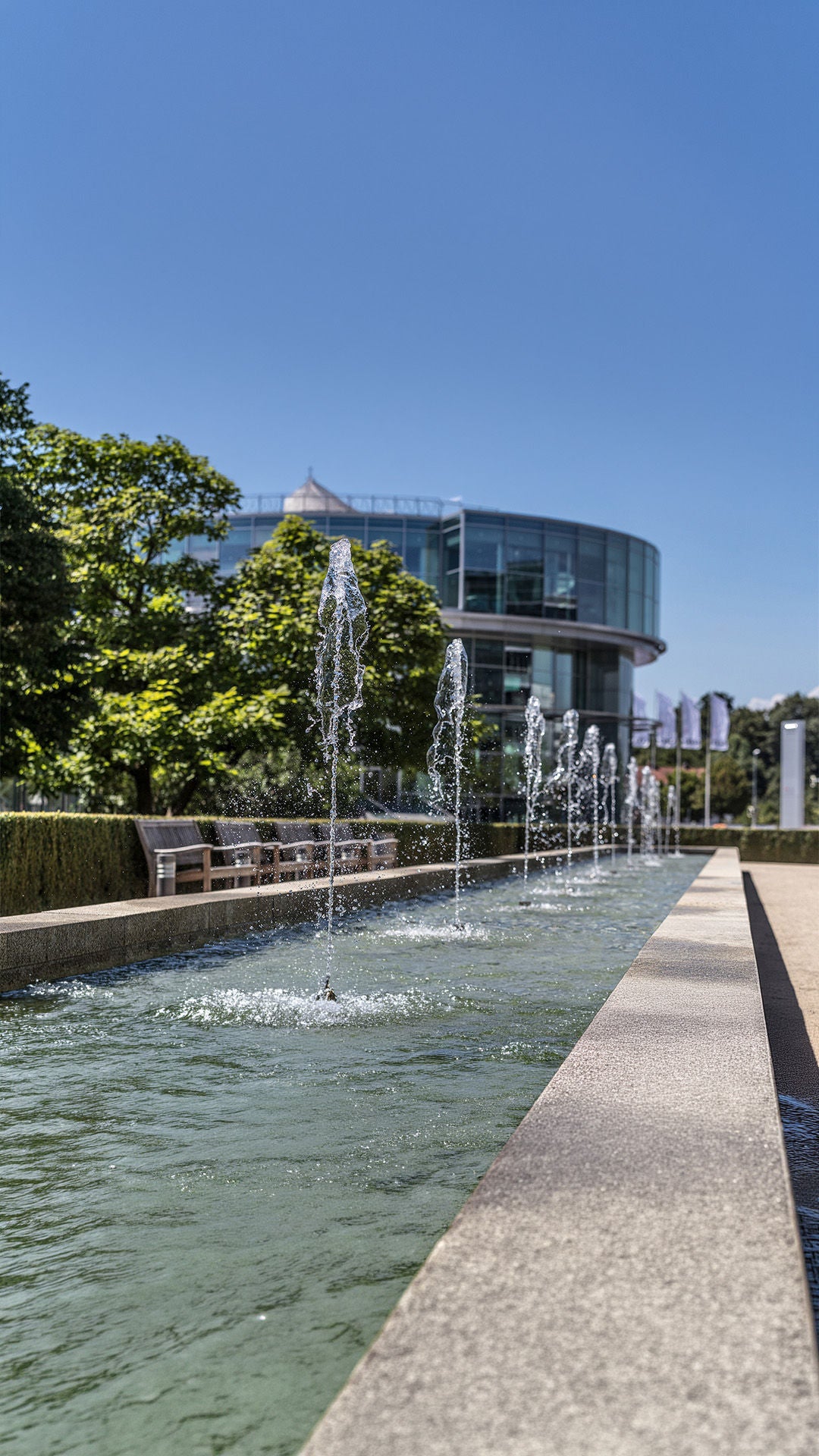 Wasserspiele in flachem Becken vor modernem Glasgebäude, Sitzbänke und Bäume am Rand.