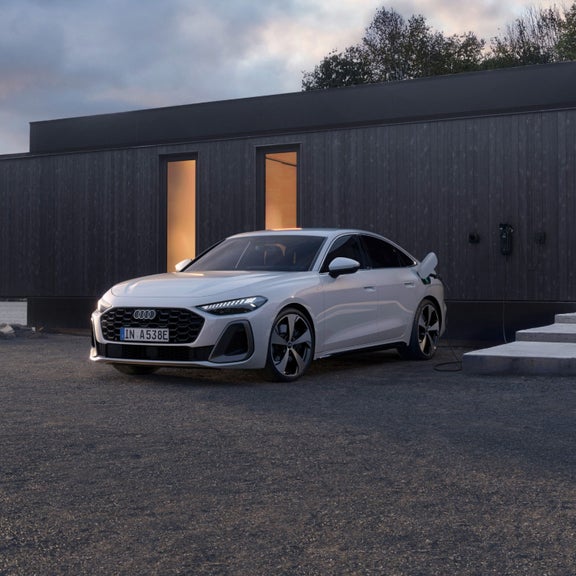 A white car is parked outside a modern, dark-clad building in the early evening. The car is plugged into a charging port, evoking a sense of modernity and sustainability.