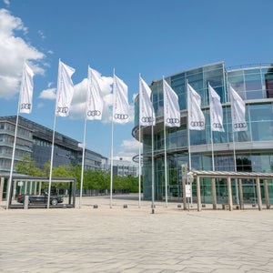 Audi Fahnen vor modernem Glasgebäude und leerem Platz bei blauem Himmel.