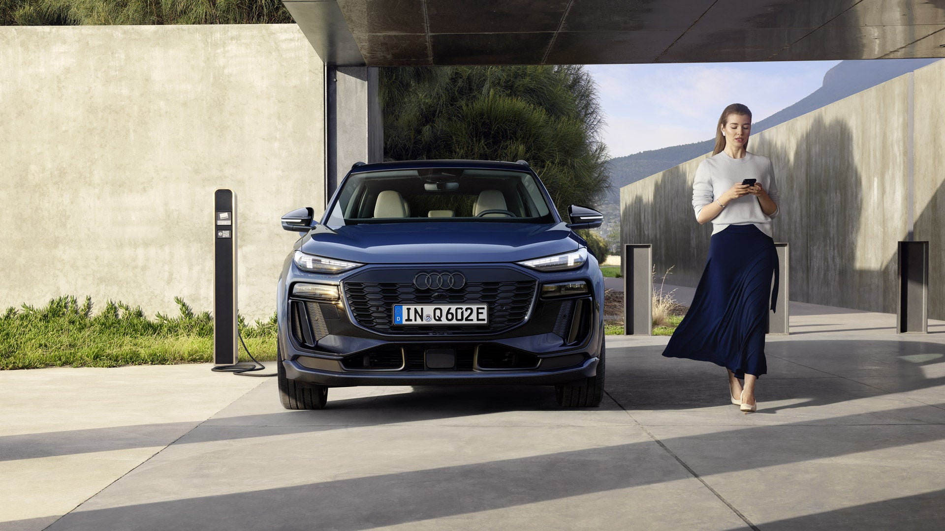Una mujer pasa junto a un auto Audi Q6 e-tron aparcado y conectado a una estación de recarga.