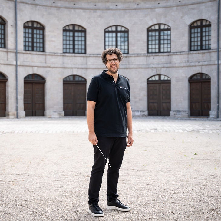 Ein Mann mit Brille, Locken und schwarzem Poloshirt steht mit Taktstock im Innenhof des gleichen Gebäudes.