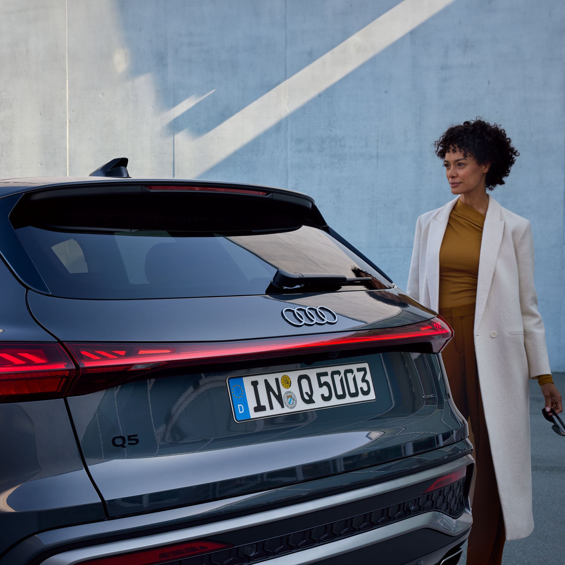 Frau in hellem Mantel steht neben einem grauen Audi Q5 vor moderner Betonwand mit Lichteffekt, Rückansicht des Fahrzeugs sichtbar.          