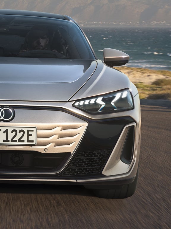 Eine frontale Nahaufnahme des Audi S e-tron GT auf einer Straße an der Küste. Die imposante Lichtsignatur der Scheinwerfer und das sportliche Design stehen im Fokus.
