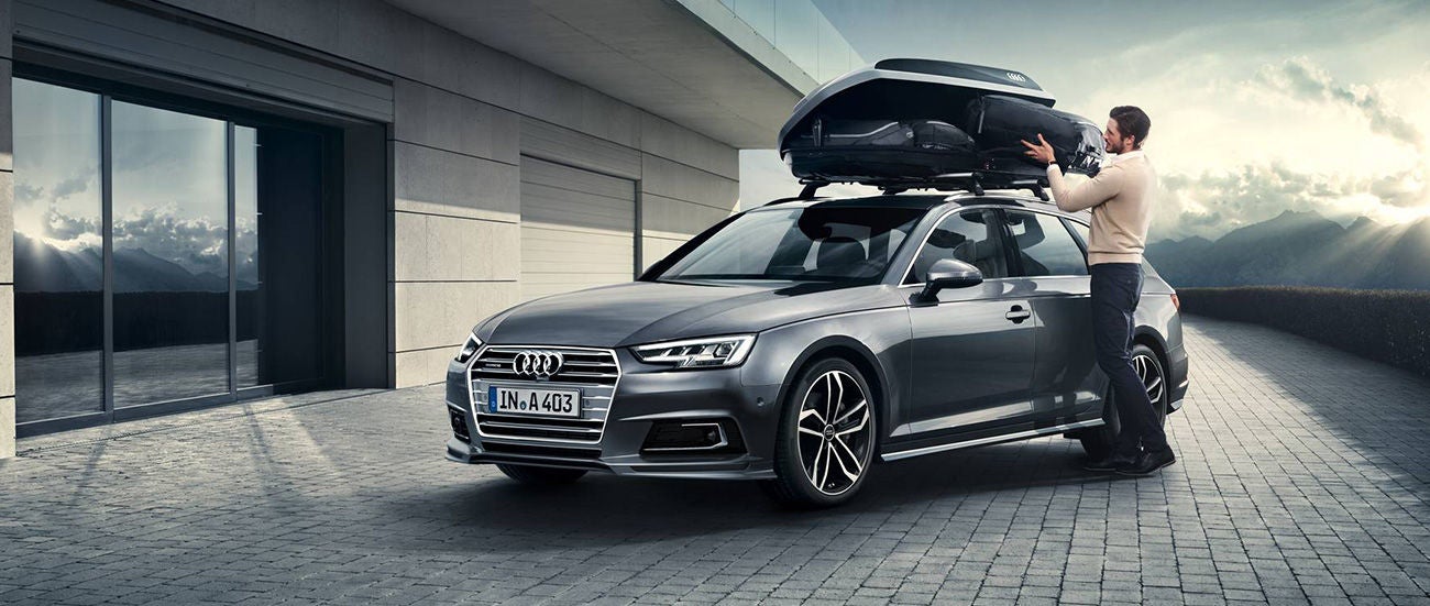Man standing next to a gray Audi with an open roof box in front of a modern building and mountain landscape.