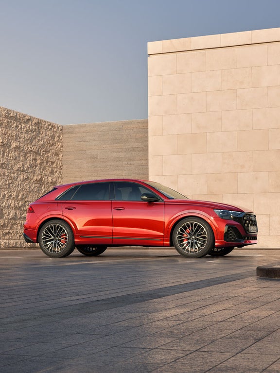 Roter Audi SQ8 SUV parkt vor moderner Steinfassade bei Tageslicht, Seitenansicht mit sportlichem Design.