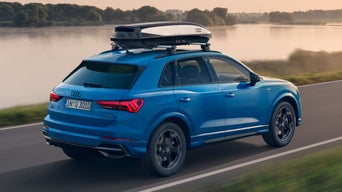 Blauer Audi Q3 mit Dachbox fährt bei Sonnenuntergang auf einer Landstraße entlang eines ruhigen Sees.