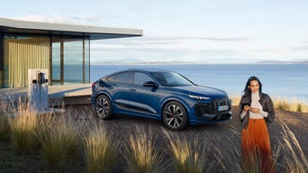 Frau mit Smartphone vor einem blauen Audi Q6 SUV e-tron, geparkt an Ladestation neben modernem Haus mit Meerblick.