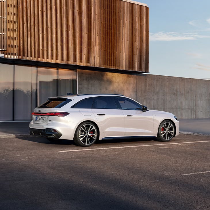 Ein Audi A5 Avant e-hybrid, der an einer Ladestation aufgeladen wird und in einer modernen städtischen Umgebung mit Gebäuden und Vegetation im Hintergrund geparkt ist.