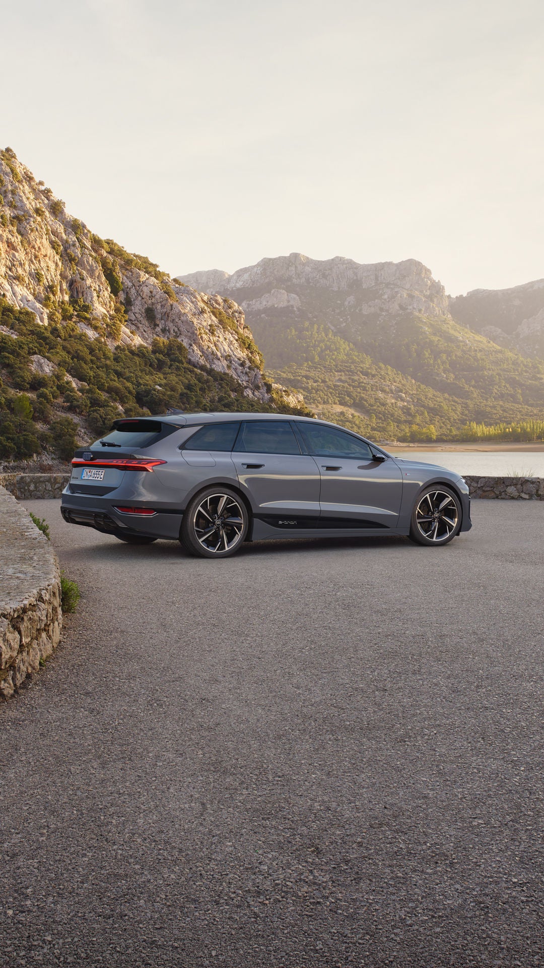 Der A6 Avant e-tron steht seitlich auf einem Kiesparkplatz vor einem See. Im Hintergrund sind von Bäumen bewachsene Berge zu sehen.