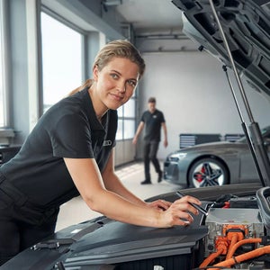 Audi Mitarbeiterin arbeitet in Werkstatt an offenem Motorraum, im Hintergrund weitere Autos. 