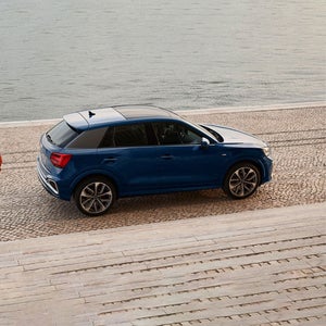 A blue SUV is parked on a cobblestone surface near a waterfront. The scene conveys a calm and serene atmosphere with gentle water in the background.