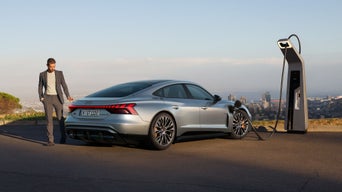 Ein silberner Audi S e-tron GT lädt an einer Ladestation mit Blick auf eine Stadt. Ein Mann im Anzug geht neben dem Auto entlang.