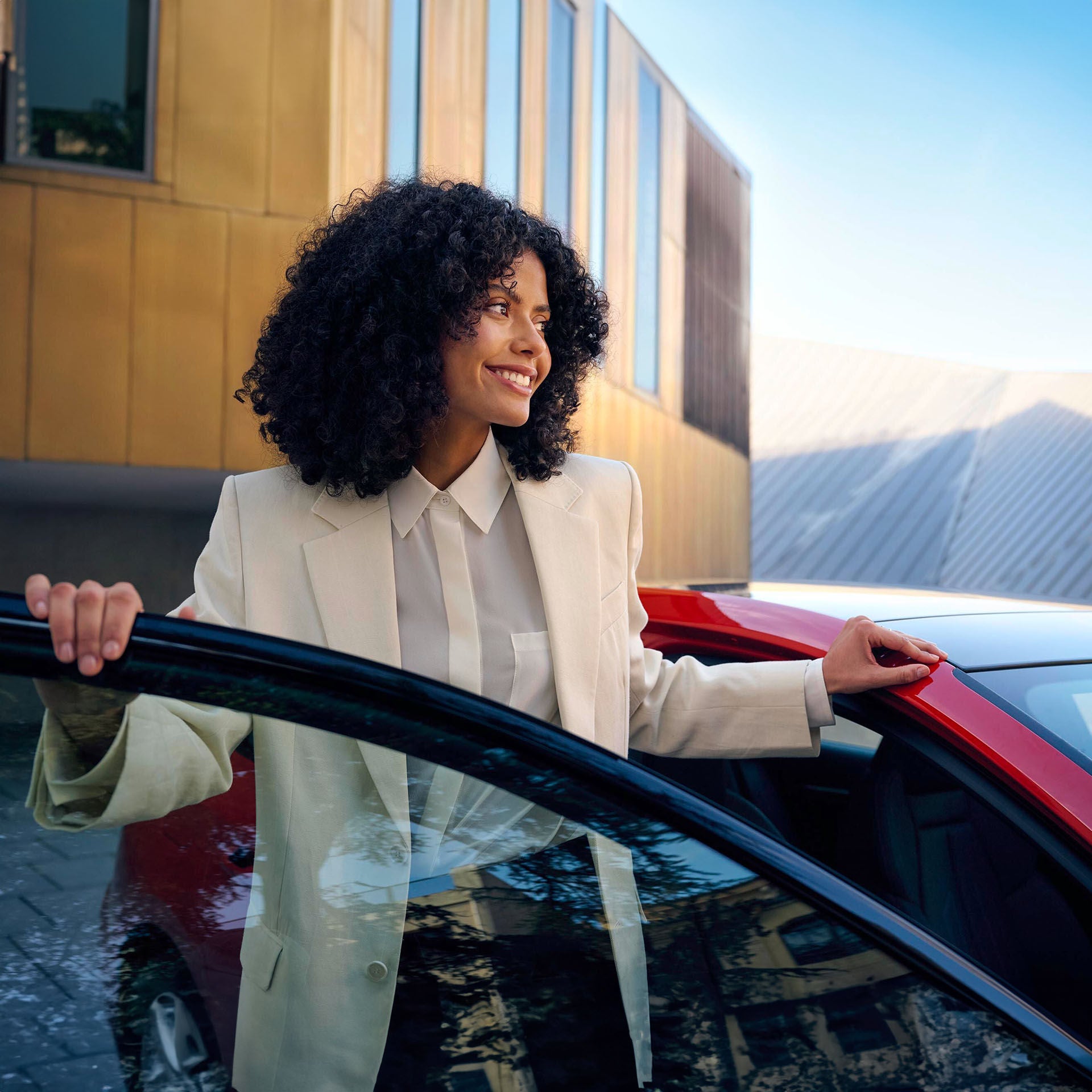 Frau in hellem Blazer steigt lächelnd aus einem roten Auto, im Hintergrund moderne Gebäude bei blauem Himmel.