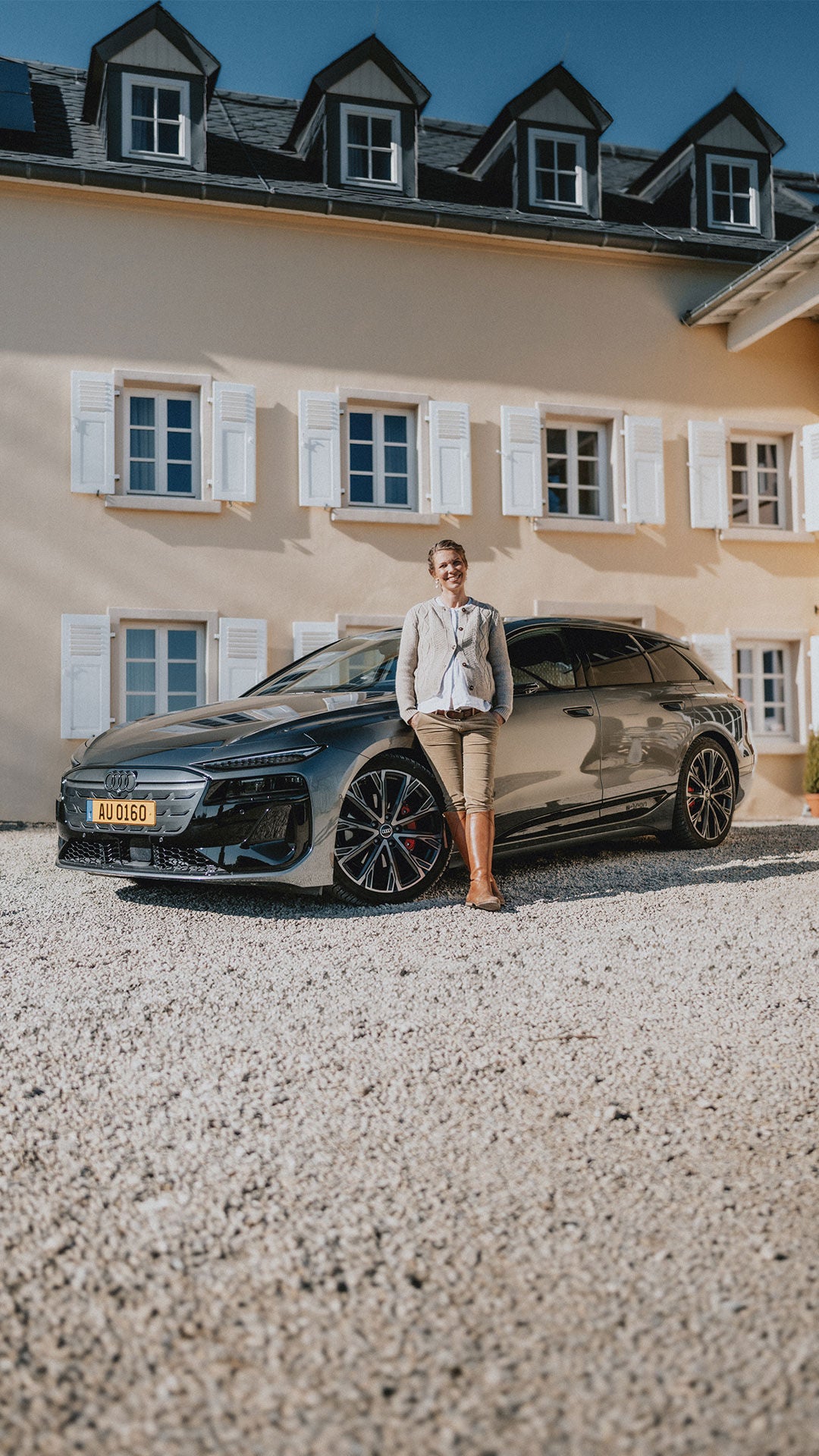 Theresa Baumgärtner lehnt an einem schwarzen A6 e-tron, der auf einem Schotterboden vor einem beigen Haus mit weißen Fensterläden steht.