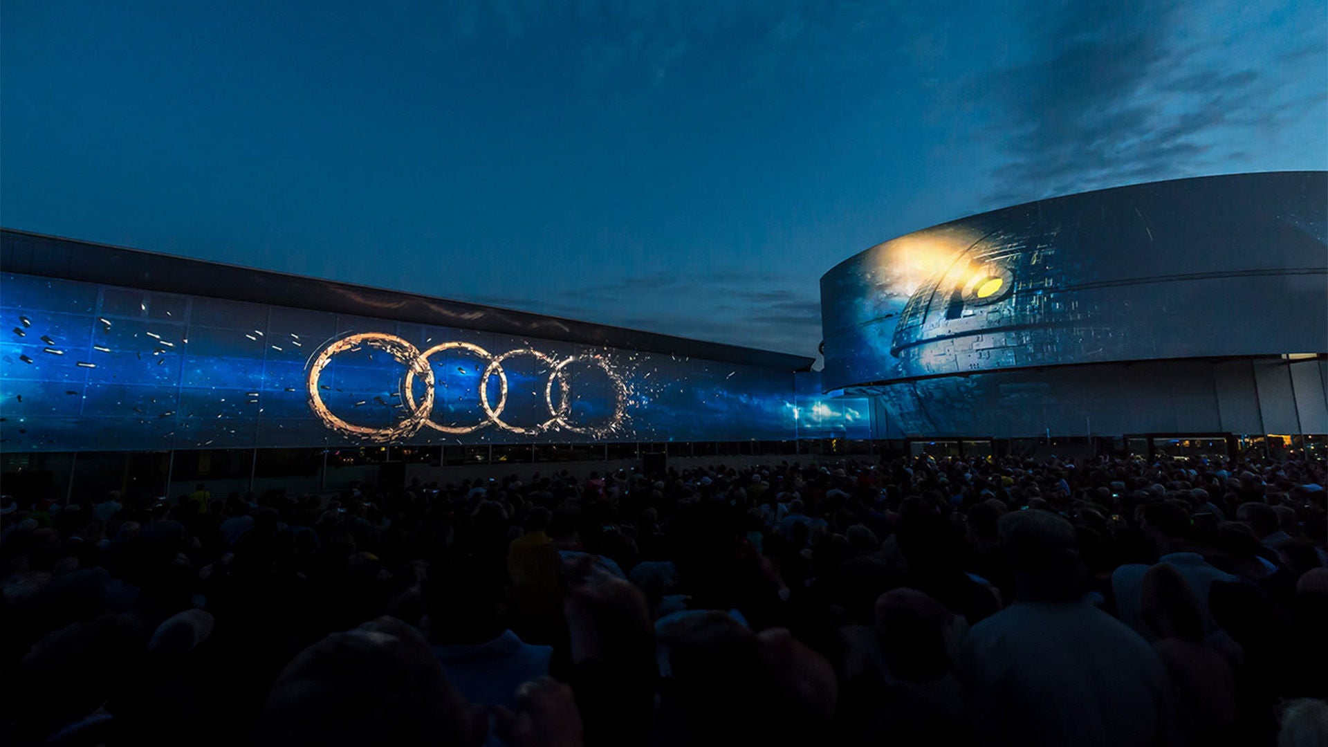 Light projection with Audi rings at night in front of a crowd and futuristic architecture.