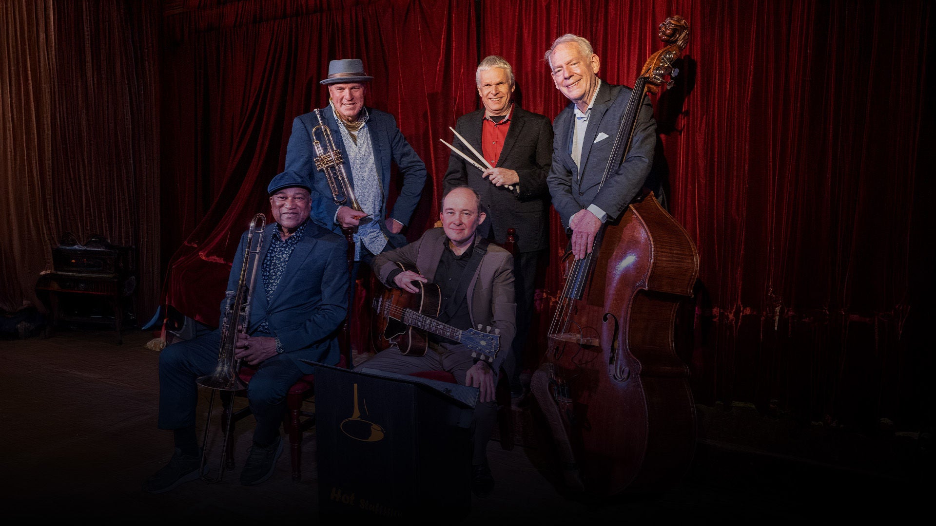 Fünfköpfige Jazzband vor rotem Samtvorhang; Musiker mit Trompete, Posaune, Gitarre, Schlagzeugstöcken und Kontrabass in gemütlicher Bühnenatmosphäre.