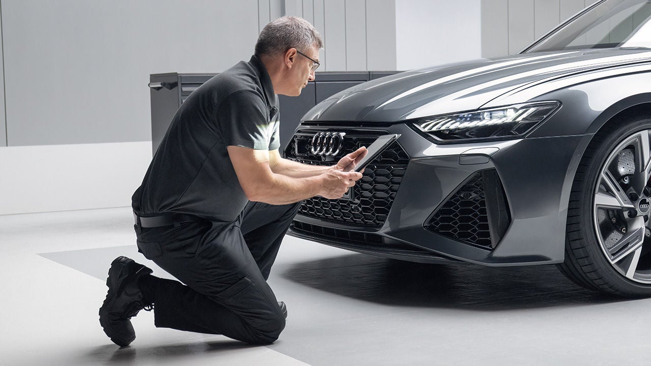Werkstattmitarbeiter kniet vor Audi-Fahrzeug und prüft mit Tablet den Frontbereich inklusive Scheinwerfer.