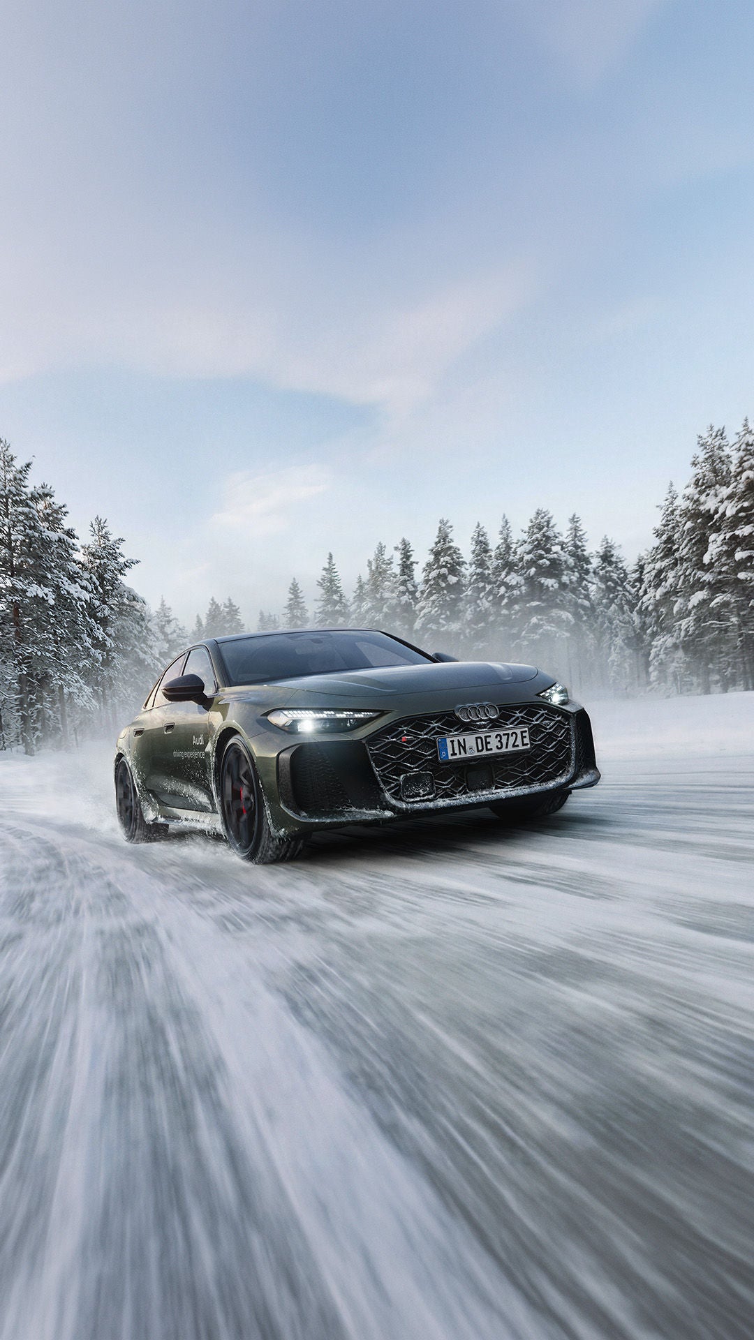 A dark green Audi RS 5 sedan drives along a snow-covered road; snow dust, a pine forest, and a blue sky.
