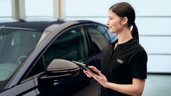 Frau im schwarzen Audi-Shirt bedient ein Tablet neben einem dunklen Auto in heller Werkstatthalle.