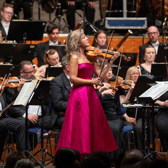 Geigerin im pinken Kleid spielt vor einem Orchester in einem festlichen Konzertsaal.