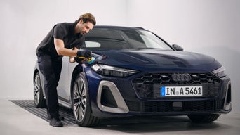 Mann poliert die Motorhaube eines blauen Audi mit Poliermaschine in heller Werkstattumgebung.