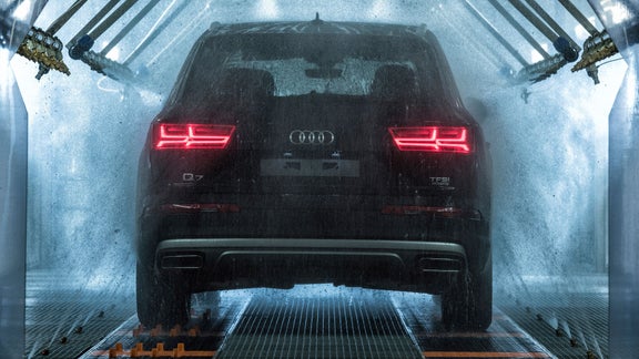 Ein schwarzer Audi Q7 wird in einer automatischen Waschanlage von Wasserstrahlen gereinigt, Rücklichter leuchten rot.