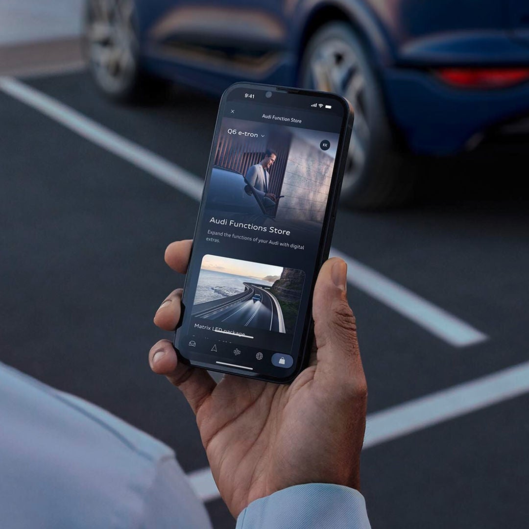 A person in a suit holds a smartphone displaying a car app, standing next to a luxury vehicle with sleek taillights.