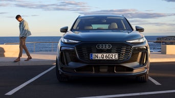 Szenische Außenaufnahme vor Küstenkulisse: Ein Audi Q6-e-tron steht frontal zur Kamera auf einem Parkplatz. Ein Mann ist im Begriff, sich vom Auto nach links zu entfernen.