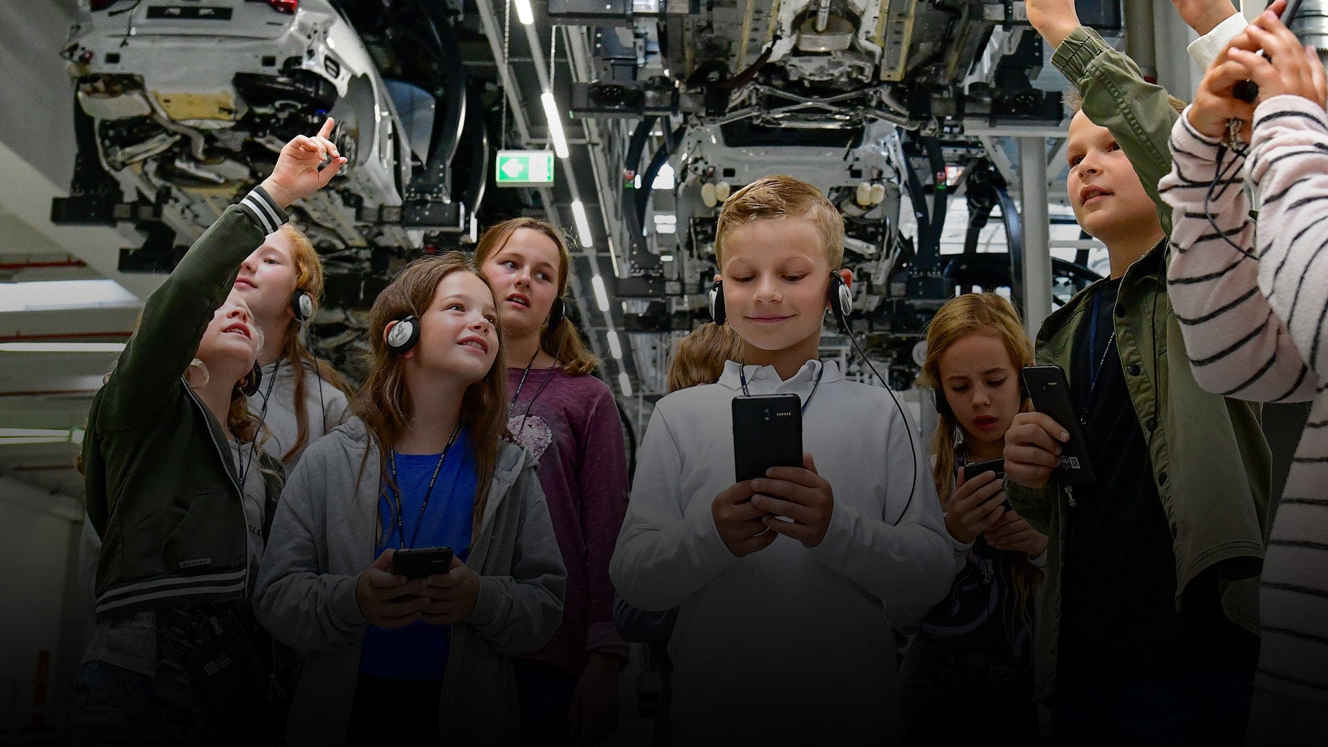 Gruppe von Kindern mit Kopfhörern bei einer Werksführung, einige zeigen auf Produktionsmaschinen.