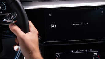Fahrer hält das Lenkrad, der zentrale Bildschirm zeigt eine Sprachsteuerungsanfrage mit der Frage: „What can I do for you?“