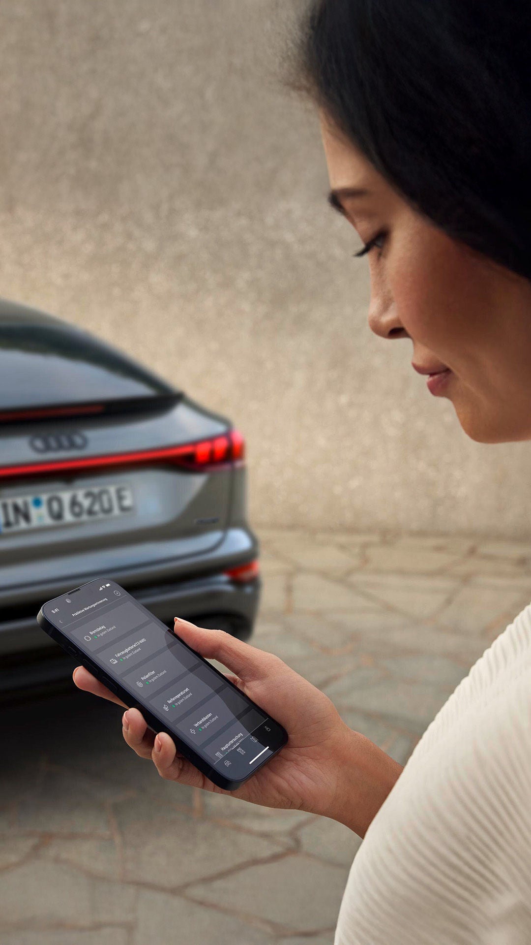 A woman looks at her phone with the myAudi app. An Audi Q6 is in the background.
