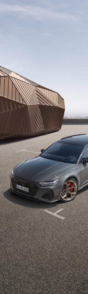 Mattgrauer Audi RS 6 Avant performance mit goldenen Felgen und roten Bremssätteln vor Gebäude mit goldfarbener Fassade auf leerem Platz.