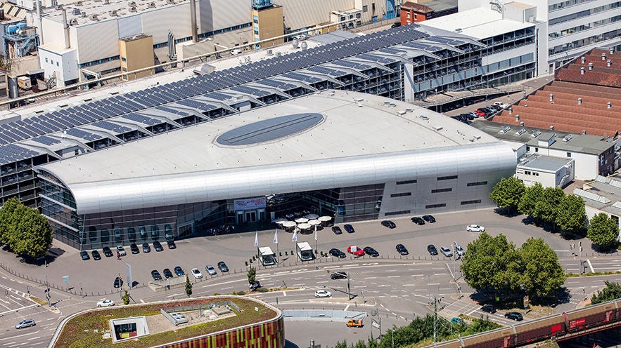Luftaufnahme eines großen Veranstaltungsgebäudes (Ankunftshalle der Audi Erlebnis Abholung in Neckarsulm) mit geschwungener Fassade, Vorplatz, Straßen und Bahntrasse.