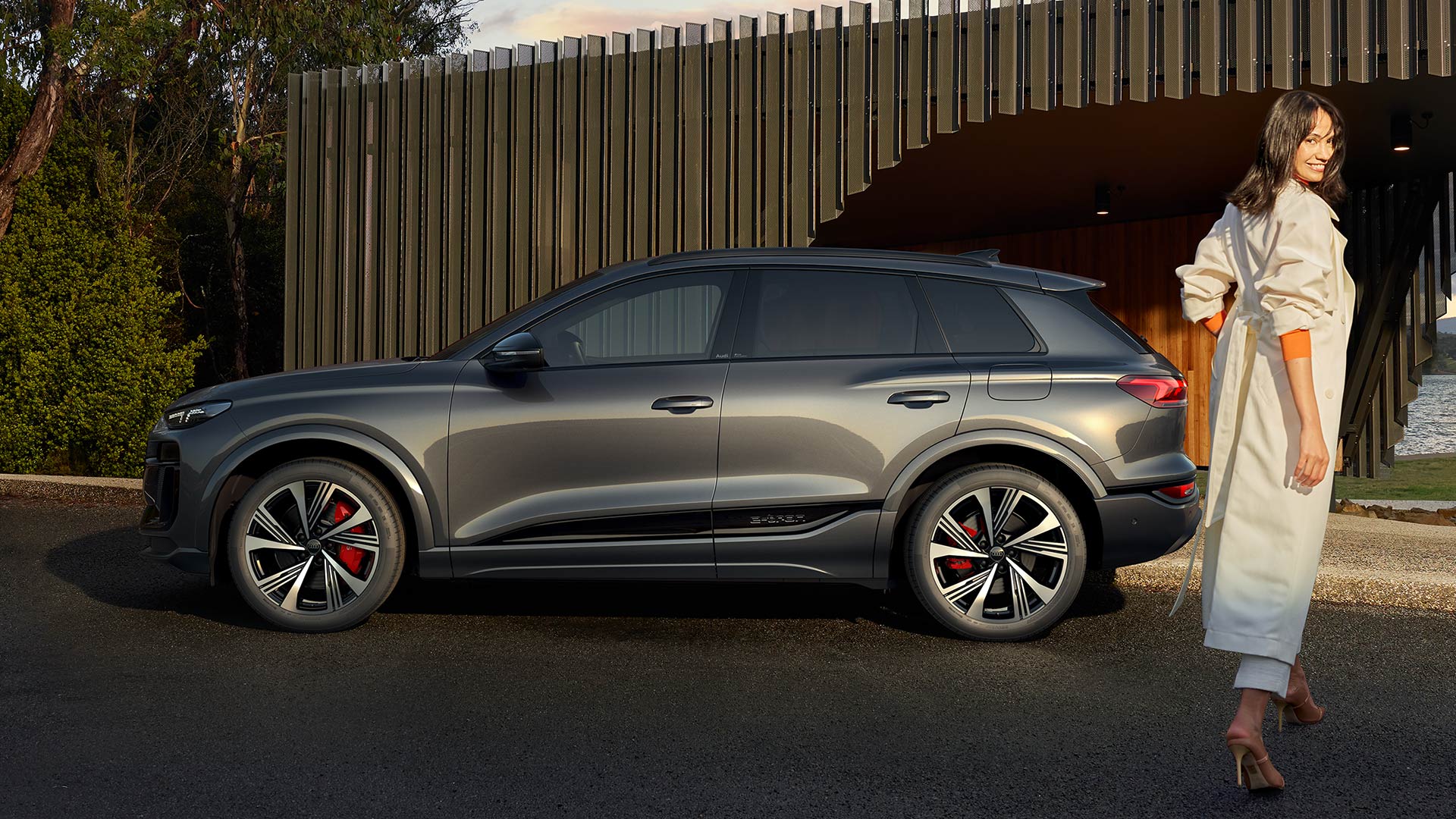 Grauer Audi SQ6 SUV e-tron seitlich vor moderner Holzfassade, eine Frau in hellem Mantel geht am Fahrzeug vorbei.