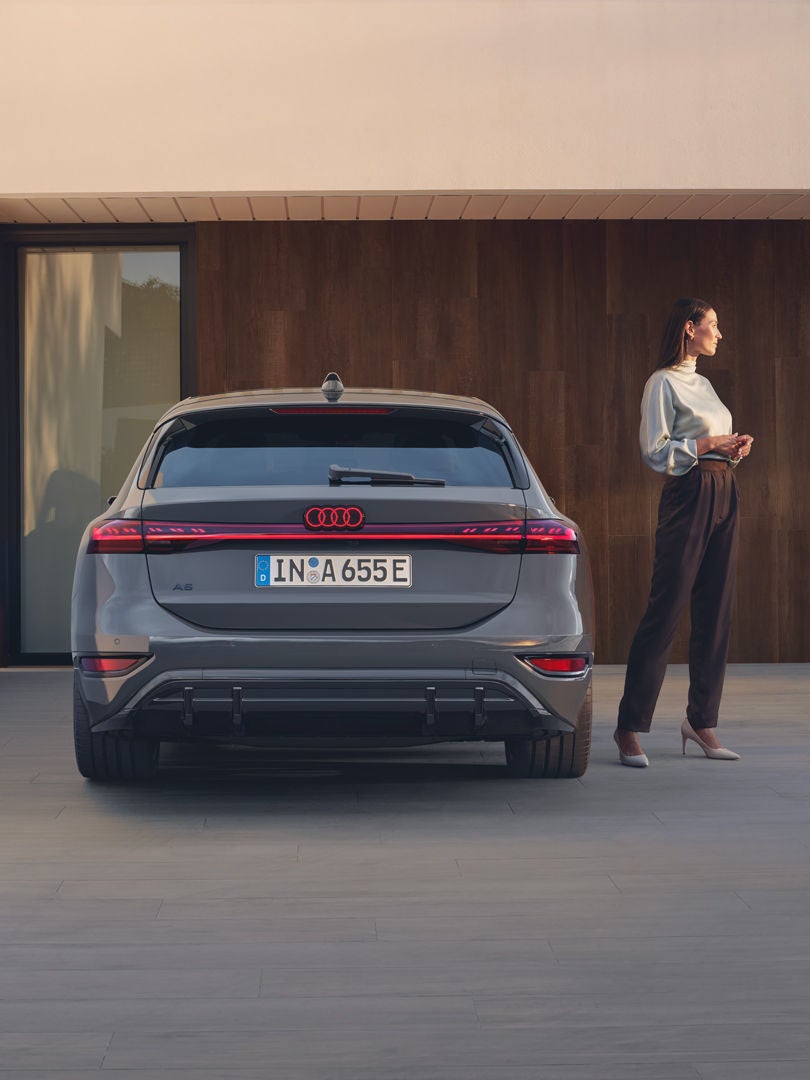 Rückansicht eines grauen Audi A6 Avant e-tron, daneben steht eine Frau vor Hausfassade.