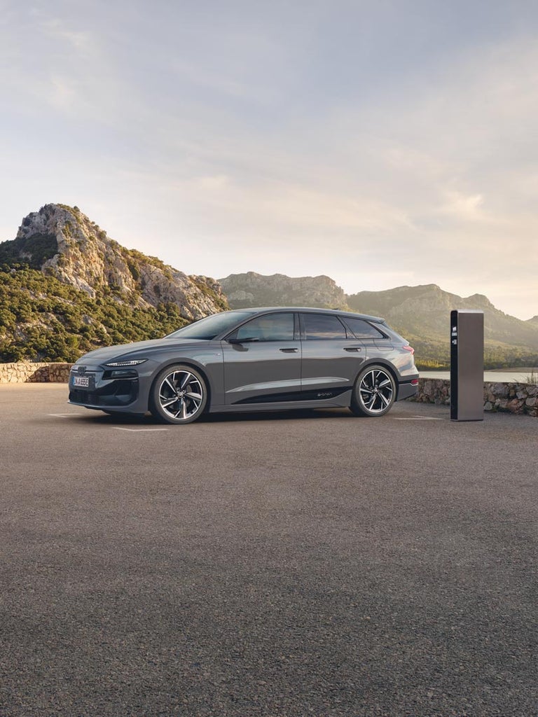 Grauer Audi A6 Avant e-tron steht seitlich auf Parkplatz vor bewaldeter Berglandschaft und Ladesäule.