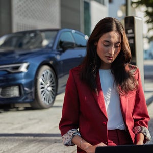 Eine Frau in rotem Hosenanzug sitzt im Freien an einem Laptop. Im Hintergrund steht ein blaues Fahrzeug vor einem Gebäude.