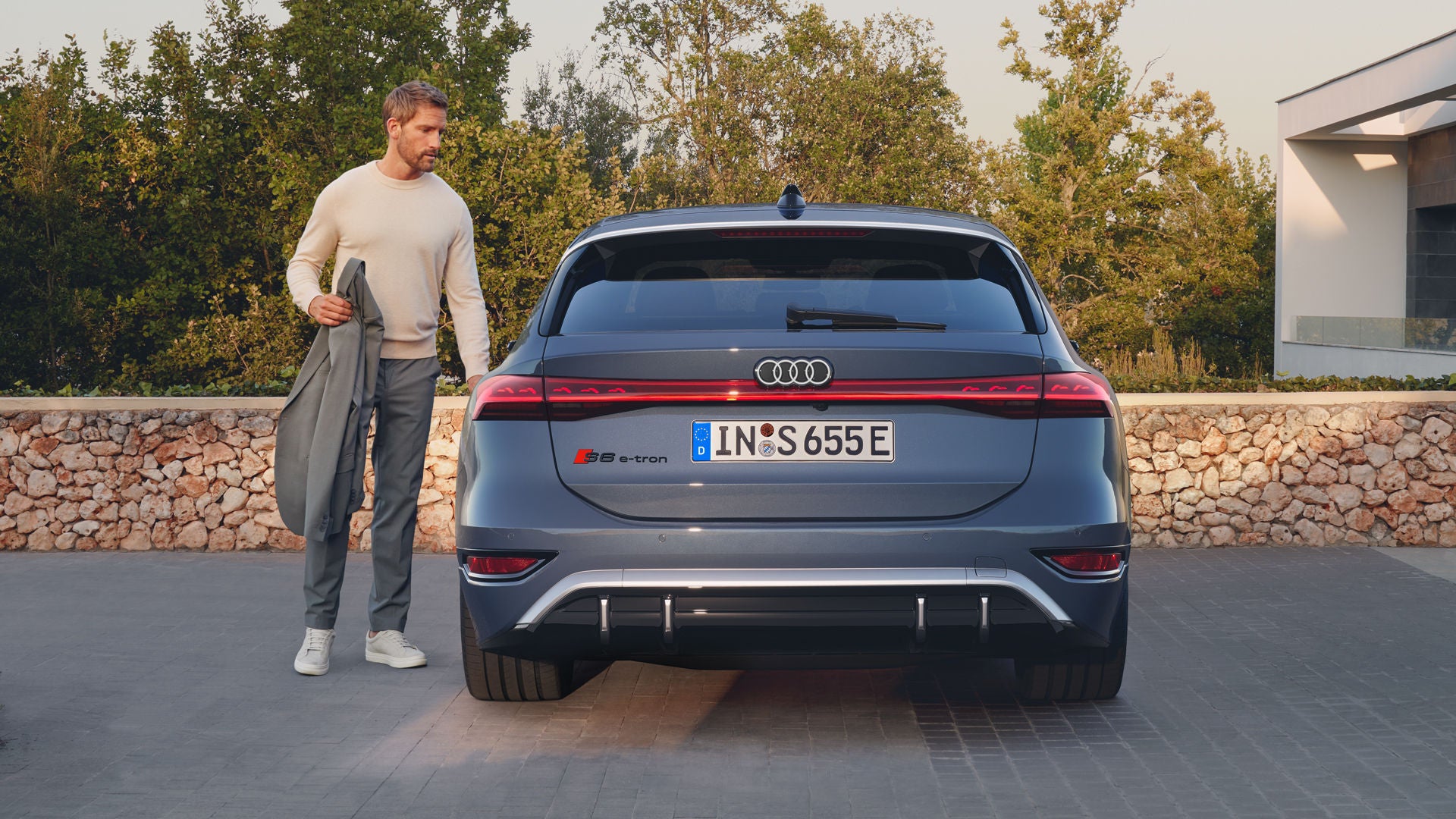 Heckprofil des S6 Avant e-tron, welcher auf Pflastersteinen vor einer Steinmauer und dem Garten eines modernen Hauses steht. Ein Mann mittleren Alters ist in Begriff, eine Tür zu öffnen.