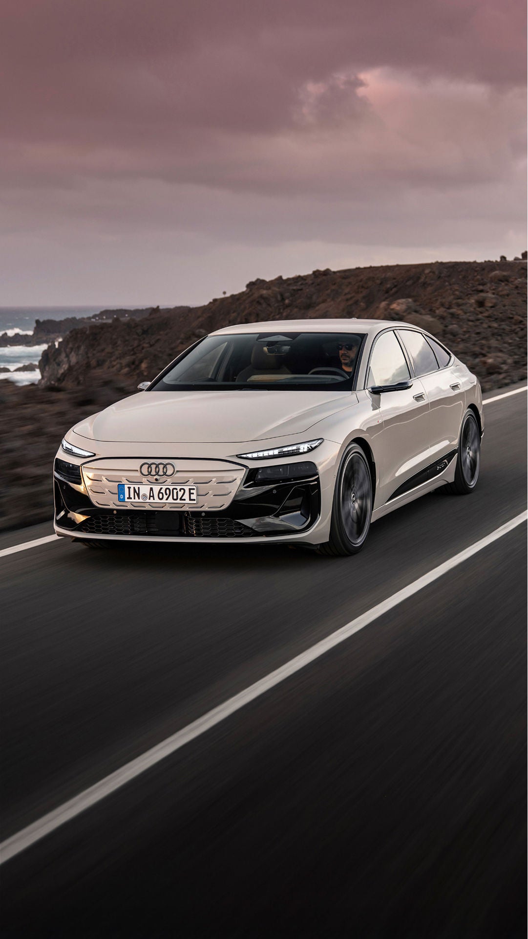 Audi sedan is driving along a coastal road, with rocky cliffs and waves crashing in the background at sunset.