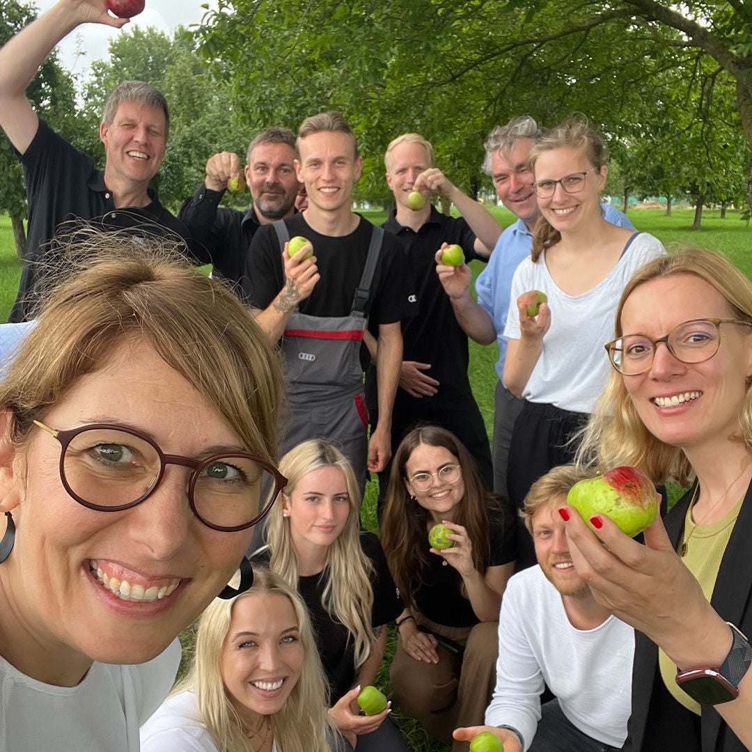 A group of cheerful people with apples in their hands stand and squat under trees on a green meadow for a photo together.