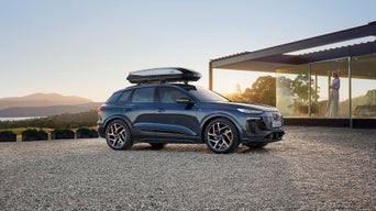 Dunkelblauer Audi Q6 e-tron mit Dachbox steht bei Sonnenaufgang vor modernem Haus, Frau blickt aus verglaster Terrasse.