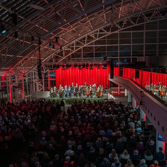 Großer Konzertsaal mit Orchester auf beleuchteter Bühne, rotem Vorhang und dicht sitzendem Publikum in festlicher Atmosphäre.