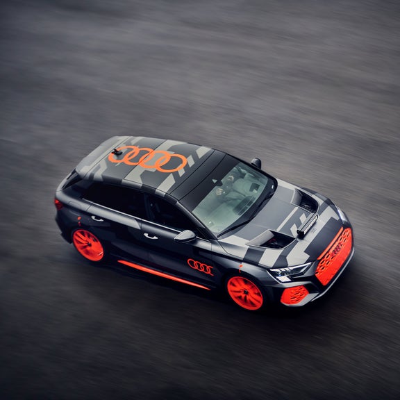 Schwarz/grauer Audi RS 3 driving experience mit orangenen Akzenten fährt auf nasser Straße, Draufsicht mit Bewegungsunschärfe. 