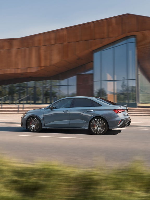 Seitliche Ansicht einer fahrenden blauen Audi RS 3 Limousine mit sportlichem Design auf einer Stadtstraße.