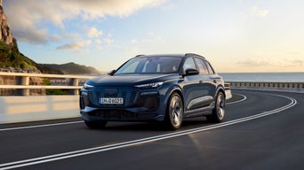 A blue Audi Q6 e-tron drives along a scenic coastal road with mountains in the background and a vibrant sky.